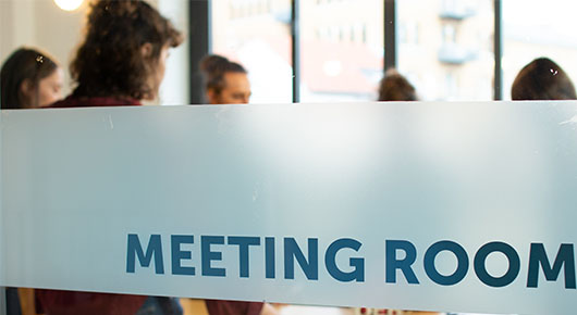 Auf einer Glaswand kann man "Meeting-Room" lesen. Dahinter sind Menschen erkennbar.